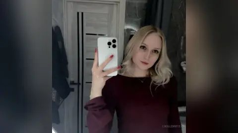 Blonde woman in a burgundy long-sleeve top taking a mirror selfie in a modern bathroom.