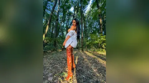 NicoleBella stands in a sunlit forest wearing a white off-the-shoulder top and thigh-high orange patterned boots.