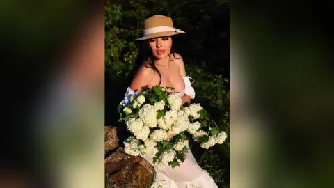 Elegant woman in a white summer dress and straw hat holding a large bouquet of white flowers in a lush garden.