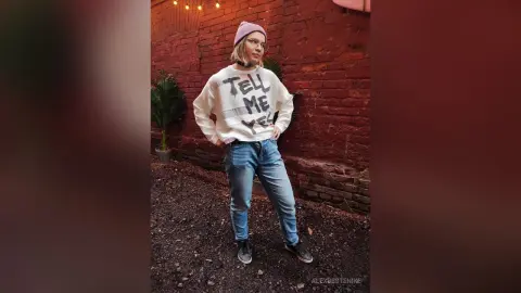 Young woman in a graphic white sweatshirt, blue jeans, and pink beanie posing confidently against a brick wall.