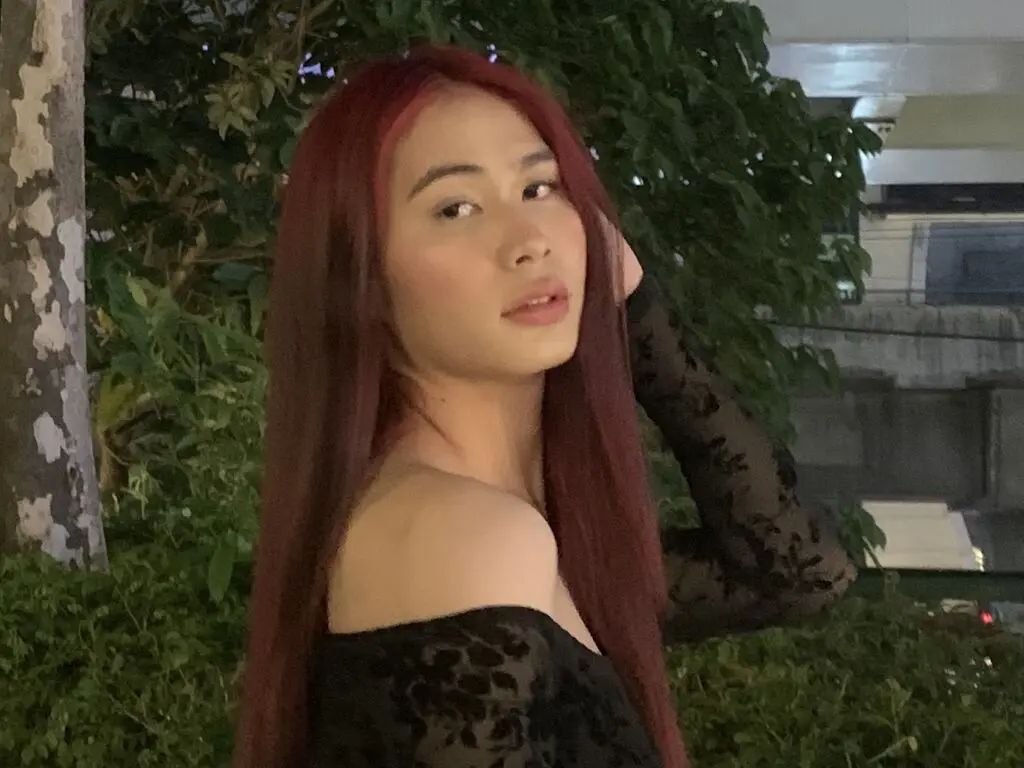 Young woman with long red hair wearing a black lace off-the-shoulder top in a lush garden setting.
