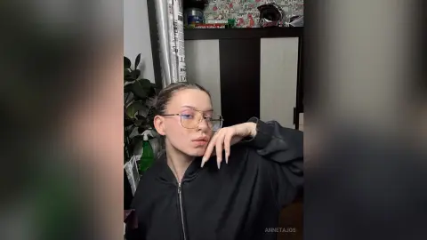 Young woman with glasses in a black zip-up hoodie posing in a cozy indoor setting.