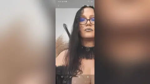 Woman with long dark hair and glasses wearing a black gothic choker in a moody indoor setting.