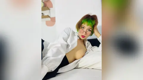 Young woman with green hair in a white button-down shirt relaxing on a white bed in a bright room.