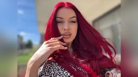 AriaVex poses outdoors with long red hair, wearing a black and white patterned top and touching her bottom lip.