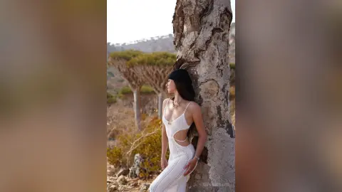 Woman in a white cutout maxi dress leaning against a tree in a scenic, sunlit landscape.