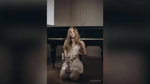 StelaBrown kneels behind a black piano wearing a white lace teddy and sheer white tights.