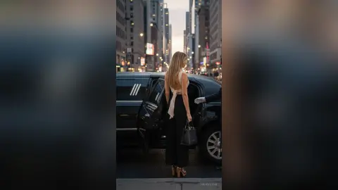 Elegant woman in a white backless top and black trousers stepping out of a luxury limousine in a city at dusk.