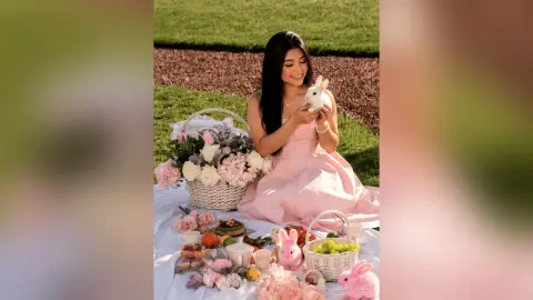 Smiling woman in a pink dress enjoying a spring picnic with flowers and bunny decorations in a sunny garden.