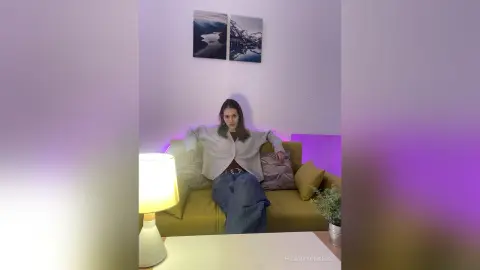 Young woman in a cozy grey sweater and jeans relaxing on a sofa in a modern, dimly lit living room.
