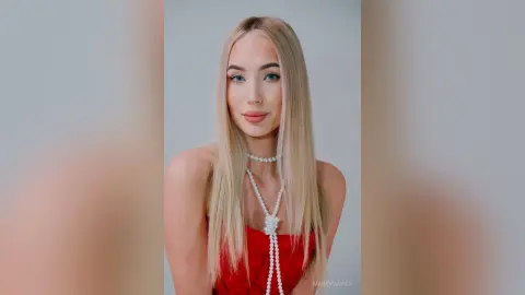 MarryDavies poses with long blonde hair wearing a red top and a white pearl choker with a long pearl necklace.
