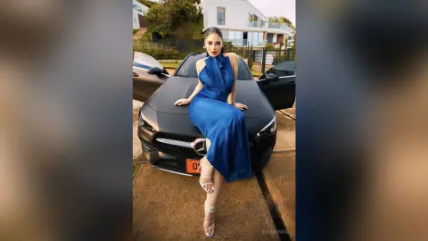 Elegant woman in a navy blue evening gown sitting on a black Mercedes luxury car in a sunny driveway.