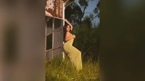 Elegant woman in a flowing yellow gown posing by a rustic white building in a dreamy meadow setting.