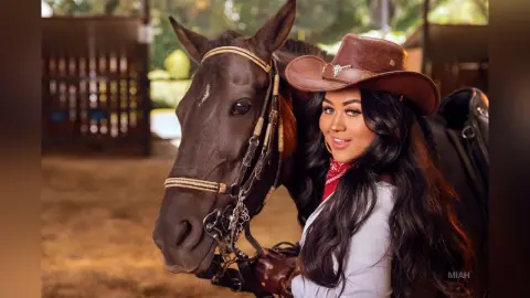 Smiling brunette woman in a cowboy hat and white shirt posing with a brown horse at a sunny ranch.
