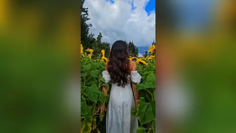 Woman in a white summer dress standing in a sunny sunflower field under a blue cloudy sky.