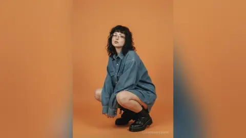 Young woman with glasses in a denim shirt and black boots posing against an orange background.