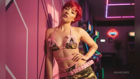 Red-haired woman in camouflage bikini leaning against a pink wall in a neon-lit retro arcade setting.