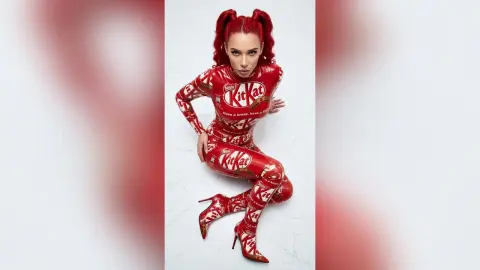 Woman in red KitKat branded bodysuit posing playfully in a bright studio setting.