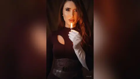 Elegant woman in a black cutout dress and white gloves holding a lit match in a moody, dark setting.