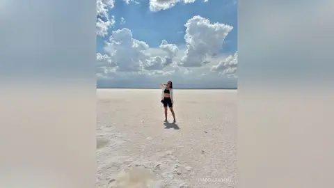 Young woman in black shorts and white shirt standing on vast white salt flats under a bright cloudy sky.