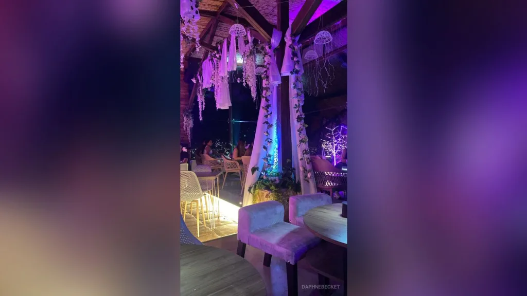 DaphneBecket — DaphneBecket sits at a table in a purple lit lounge decorated with hanging white fabrics and fairy lights.