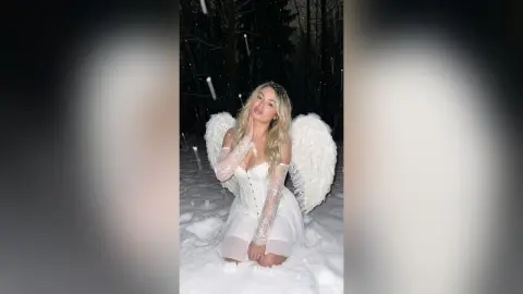 Blonde woman in a white angel costume with wings kneeling in a snowy forest at night.