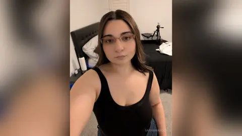 Young woman in a black tank top taking a casual selfie in a cozy bedroom setting.