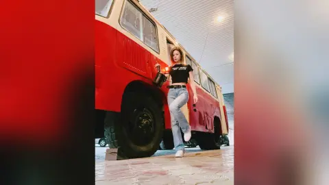 Stylish young woman in black crop top and jeans posing by a vintage red bus in a moody urban setting.