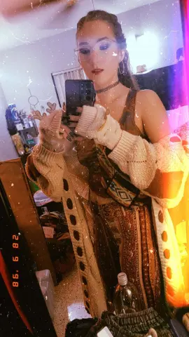 jenellehayesxoxo wears a patterned brown tank top and a chunky cream cardigan while taking a mirror selfie.