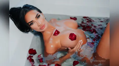 AnnaKarev is naked in a bubble bath with red rose petals scattered on her skin and in the water.