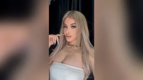 ElinaCarter poses with her long blonde hair and heavy eye makeup, wearing a white tube top and a gold necklace against a black background.