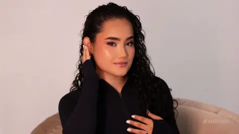 JeanRusso poses with her long, curly dark hair and heavy makeup, wearing a black long-sleeved top against a plain white background.