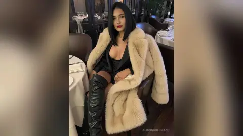 Glamorous woman in a black outfit and cream faux fur coat sitting at a fine dining restaurant table.