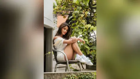 TefyRusso sits on a chair outdoors, wearing a white off-the-shoulder dress and white sneakers, posing with her legs bent and hands resting on her knees.