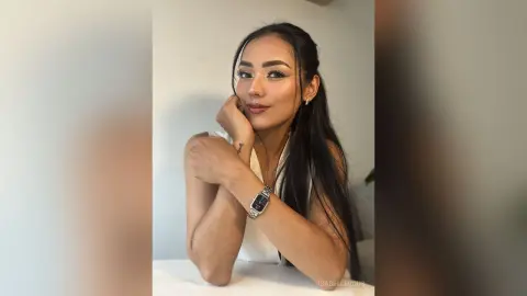 Smiling woman with long dark hair wearing a white sleeveless top in a bright, minimalist indoor setting.