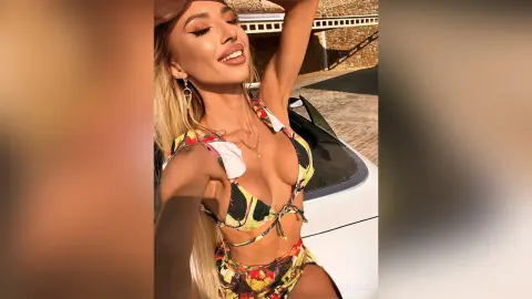 Blonde woman in colorful floral bikini posing outdoors by a white car in bright sunlight.