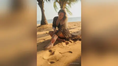 Young woman in a leopard print maxi dress sitting on a sunny tropical beach at sunset.
