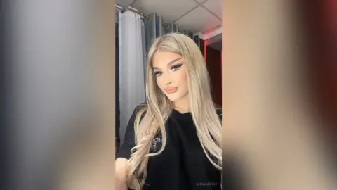 ElinaCarter poses with long blonde hair and heavy eye makeup, wearing a black t-shirt in a room with white curtains.
