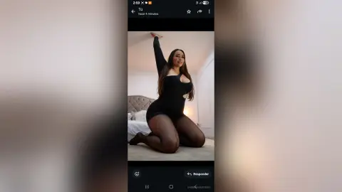SabrinaVantress poses kneeling on a floor in a black bodysuit and sheer black tights, raising one arm while looking at the camera.