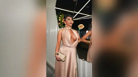 Dolce poses in a low-cut, champagne-colored satin gown that shows deep cleavage, holding a gold clutch bag.