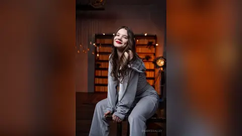 Smiling young woman in a grey lounge set sitting in a cozy, dimly lit room with warm ambient lighting.