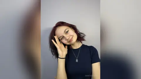 Smiling young woman with reddish hair wearing a black t-shirt and silver necklace against a plain gray wall.