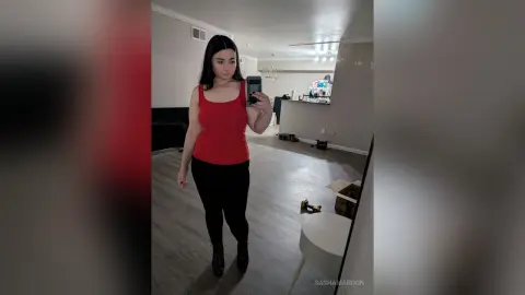 SashaMaroon stands in a room wearing a red tank top and black leggings while taking a mirror selfie.