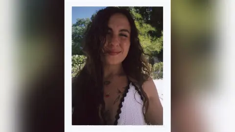 Smiling woman with long wavy hair wearing a white crochet summer top in a sunny outdoor garden setting.