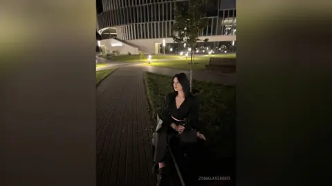 Young woman in a black long-sleeve top sitting on a park bench at night near a modern city building.