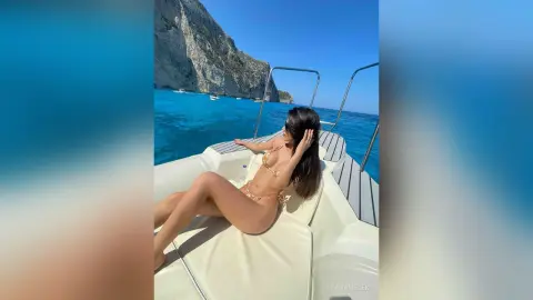Young woman in a bikini relaxing on a white boat in turquoise ocean waters near rocky cliffs.