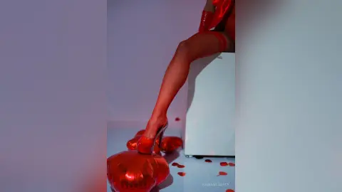 Elegant woman in a red outfit and high heels sitting on a white pedestal in a minimalist studio setting.