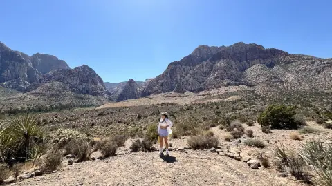 Adventurous woman in white summer outfit standing in a sunny desert landscape with rocky mountains.