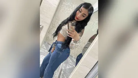 Young woman with long dark hair in a floral crop top and blue jeans taking a mirror selfie in a bright bathroom.
