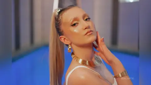 Glamorous woman with gold glitter makeup and jewelry posing in a blue lit setting.